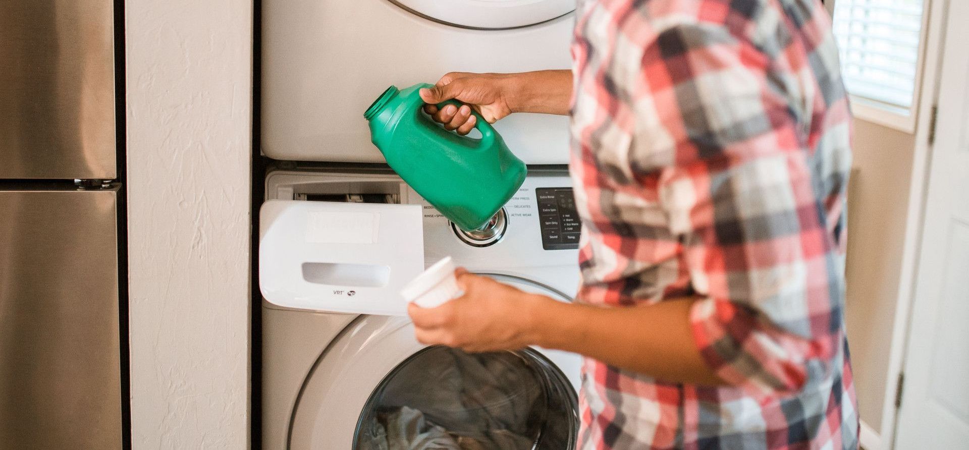 A man in a plaid shirt pours detergent into a front-loading washing machine, engaging in a household chore.